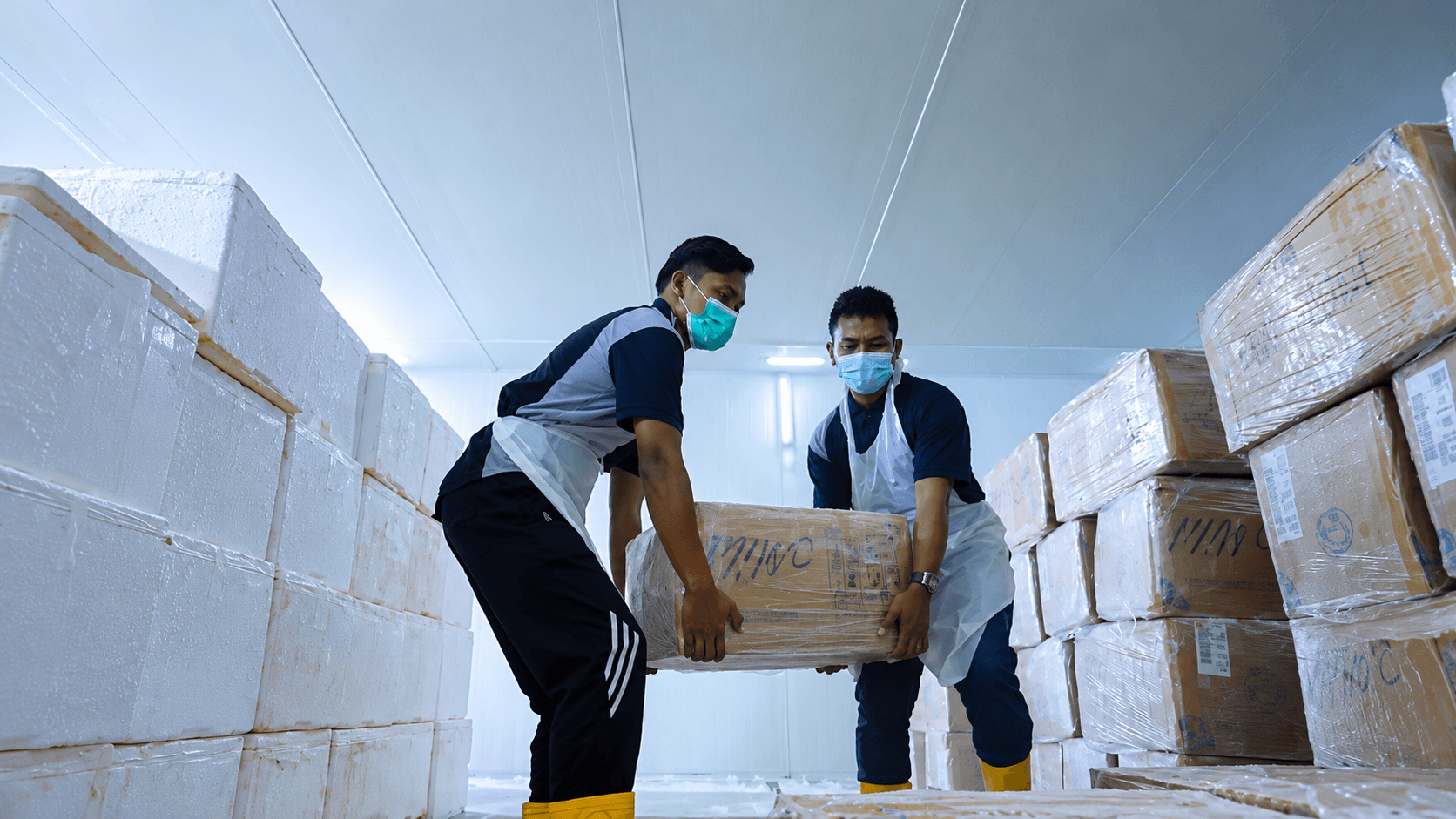 Two workers wearing face masks carry a cardboard box inside a chilled-looking storage room.