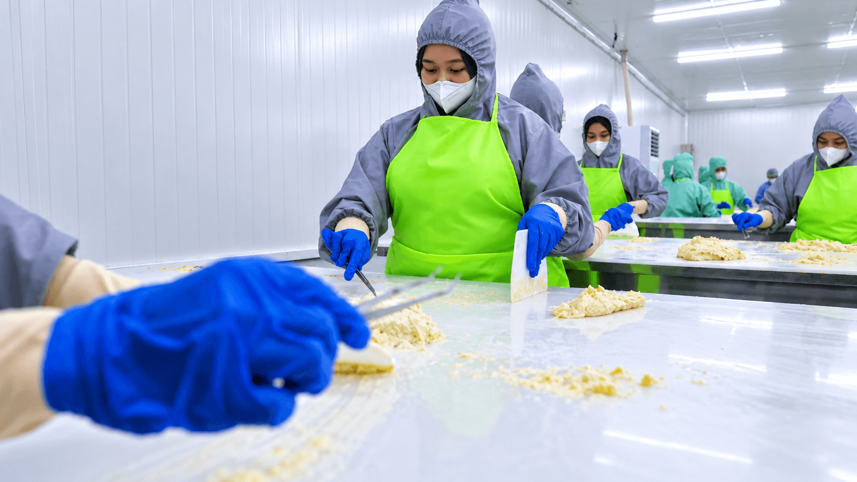 Workers in protective clothing and gloves handle durian flesh on stainless-steel tables in a food processing room.