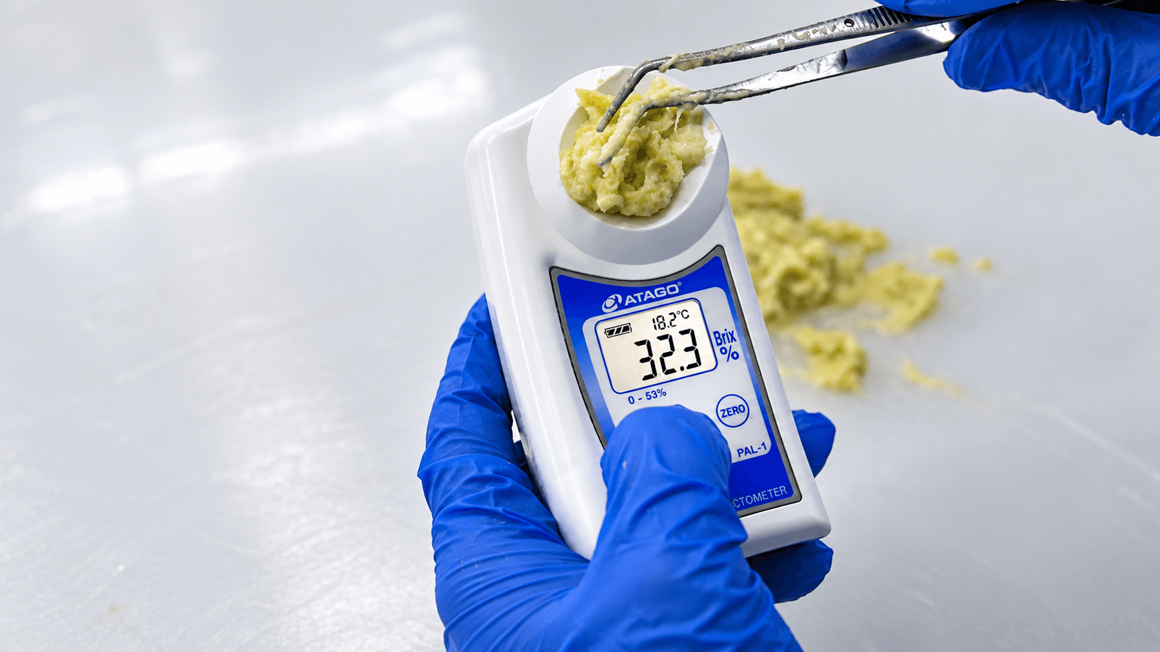 Blue-gloved technician measuring a sample of durian pulp with a handheld digital refractometer.
